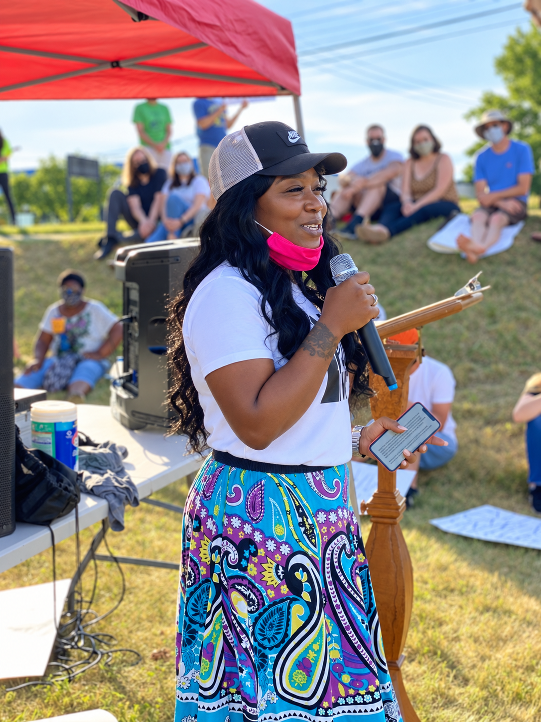 Marsita speaking into a microphone at a Juneteenth rally, wearing a white Black Lives Matter shirt and a colorful skirt